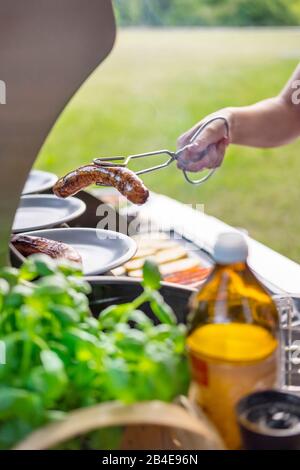 Hand hält Grillzangen mit Grillwurst, Grill Stockfoto