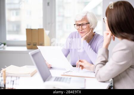 Frau im Alter, die während eines Meetings im Büro am Schreibtisch neben dem jungen Agenten sitzt und das Versicherungsformular durchschaut Stockfoto