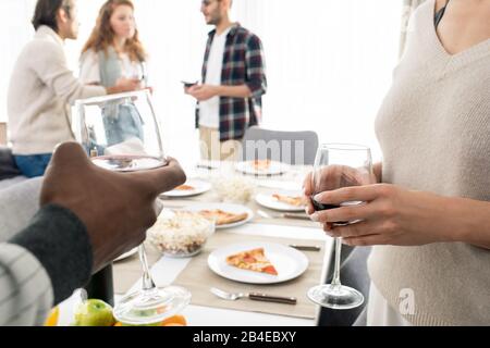 Hände des jungen interkulturellen Paares mit Brille von Rotwein, das zu Hause auf dem Hintergrund des servierten Tisches und der Freunde toastert Stockfoto