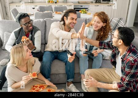 Zwei glückliche junge Männer, die sich gegenseitig fünf geben, während sie auf der Couch unter Freunden sitzen, die auf der Heimparty sitzen Stockfoto