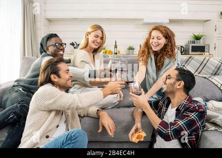 Fröhliche junge multikulturelle Freunde, die mit Brille Rotwein klinken, während sie auf der Heimparty auf der Couch toasten Stockfoto