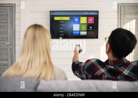 Junger Mann mit Fernbedienung, der etwas zu sehen hat, während er mit seiner Freundin in der Nähe vor dem fernseher sitzt Stockfoto