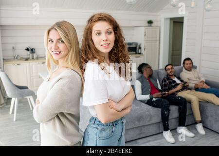 Zwei hübsche Mädchen mit gekreuzten Armen auf der Brust stehen vor der Kamera, wobei sich eine Gruppe von Jungs auf der Couch auf dem Hintergrund entspannen Stockfoto