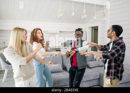 Zwei junge interkulturelle Paare tanzen gemeinsam im Wohnzimmer zu Hause auf dem Hintergrund von Couch und Küche Stockfoto