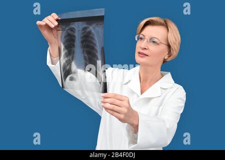 Selbstbewusster Arzt im mittleren Alter, isoliert auf blauem Hintergrund stehend, Röntgenbild in den Händen halten, Lungen überprüfen und ruhig lächeln Stockfoto