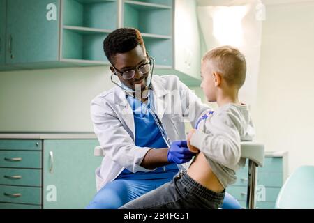 Arzt, der den Patienten betrachtet und die Haltung des kleinen Jungen untersucht. Fröhliches Kind lächelt und schaut weg. Afroamerikanischer Arzt mit Stethoskop am Hals, Stockfoto