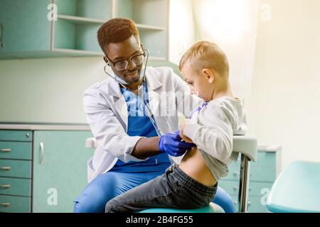 Arzt, der den Patienten betrachtet und die Haltung des kleinen Jungen untersucht. Fröhliches Kind lächelt und schaut weg. Afroamerikanischer Arzt mit Stethoskop am Hals, Stockfoto