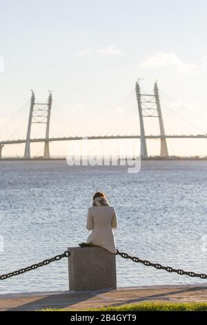 Einsamkeit, der Wunsch, allein zu sein, spazieren Sie am Wasser entlang. Frau, die auf dem Meer sitzt und die Brücke betrachtet. Ansicht von hinten. Stockfoto