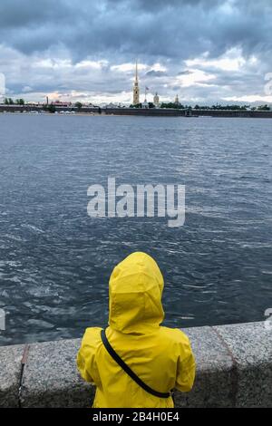 Rückansicht der einsamen Frau in gelbem Regenmantel und Kapuze, die auf dem Böschung steht und auf den Fluss Newa in St. Petersburg blickt. Übertrieben depressiv Stockfoto