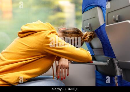 Frau in orangefarbenem Hoodie, die auf einem Sitz sitzt und in öffentlichen Verkehrsmitteln auf einem Klapptisch schläft. Müde erschöpfte junge Frau nach der Arbeit schlief in t ein Stockfoto