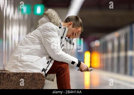 Weinender Mann in weißer Parka sehr verärgert, hält Smartphone, bekommt schlechte Nachrichten, verhüllt sein Gesicht mit der Hand, sitzt auf der Bank in der U-Bahn-Station. Problem in Stockfoto