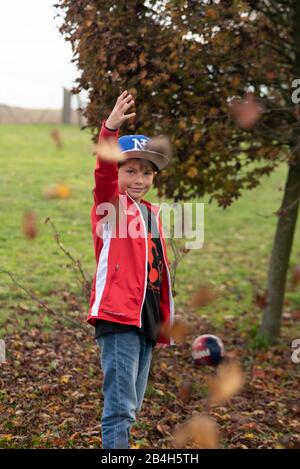 Ein achtjähriger Junge wirft Blätter in die Luft, er freut sich auf den Herbst und das nächste Fußballspiel, im Hintergrund steht sein Fußball vom französischen Verein Paris Saint Germain, Stockfoto