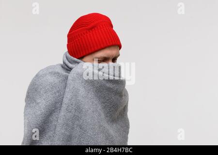 Kranker Mann mit roter Mütze, in warmgrauen Pflaumen gehüllt, an Kälte, Virus, Fieber, Blick nach unten, isoliert auf grauem Hintergrund. Grippesaison. Stockfoto