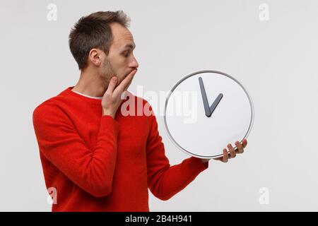 Mann in rotem Pullover mit großer rund-um-Uhr, bei Schock bedeckt er seinen Mund mit der Hand. Zeitmangel, physische Überarbeit, Terminversetzung, Zeitverlastung Stockfoto