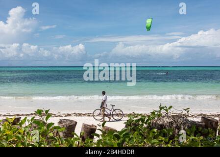 Sansibar, Tansania: Traumstrand an der Ostküste dieser afrikanischen Insel im Indischen Ozean. Islander schiebt sein Fahrrad am Strand, Kitesurfer im Hintergrund. Stockfoto