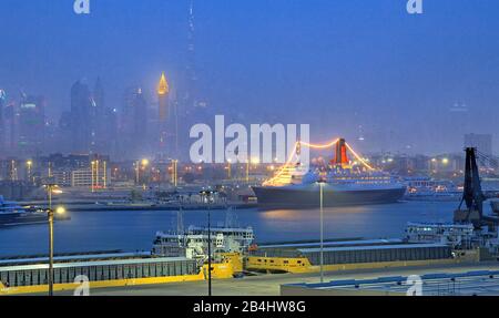 Hotel- und Museumsschiff Queen Elizabeth 2 (QE2) im Hafen mit Stadtsilhouette und Burj Khalifa 828m nachts, Dubai, Persischer Golf, Vereinigte Arabische Emirate Stockfoto