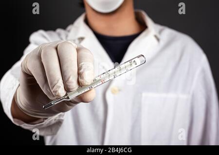 Doktor in einem weißen Mantel in einer Maske und Handschuhe hält ein Thermometer in der Hand. Behandlung auf virale und bakterielle Krankheit prüfen.Körpertemperatur erhöht f Stockfoto