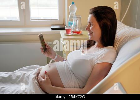 Hochriskige Schwangerschaft, schwangere Frau, die im Krankenhaus auf dem Mutterschaftsbett liegt und auf ihrem Handy sucht Stockfoto