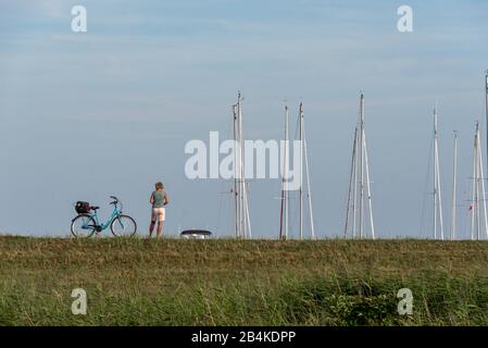 Deutschland, Mecklenburg-Vorpommern, Vitte, eine Frau steht auf einem Deich auf der Insel Hiddensee und schaut auf ihr Handy. Neben ihr befindet sich ein Fahrrad. Stockfoto