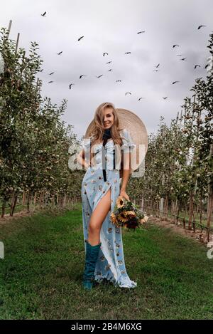 Blonde Mädchen in einem blauen Kleid stehen zwischen dem Apfelgarten, Sommer Blumen in ihren Händen Stockfoto