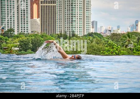 Ein Mann schwimmt einen Schmetterling in einem Pool auf dem Dach eines Luxushotels. Blick auf die Stadt Manila vom Pool des luxuriösen Fünf-Sterne-Discovery Primea Hot Stockfoto