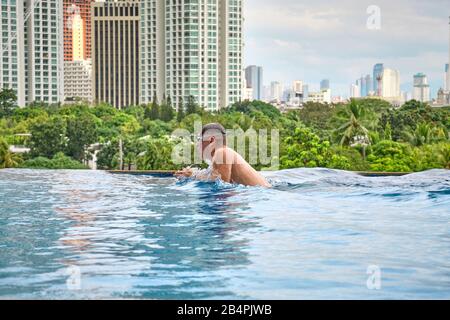 Ein Mann schwimmt einen Schmetterling in einem Pool auf dem Dach eines Luxushotels. Blick auf die Stadt Manila vom Pool des luxuriösen Fünf-Sterne-Discovery Primea Hot Stockfoto