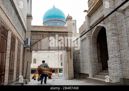 Mann, der Stapel Besen zum Verkauf in der Seitenstraße in der Altstadt von Buchara, Usbekistan, trägt Stockfoto