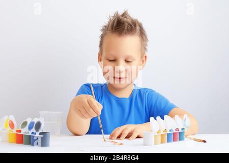 Fröhlicher fröhlicher kleiner Junge mit unordentlichen Haaren hält einen Pinsel in der Hand und zieht mit goldener Farbe. Bildung in der Schule oder im Kindergarten. Stockfoto