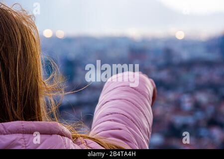 Mädchen mit braunem Haar und rosafarbener Jacke, die in der Ferne in Richtung Stadt zeigt Stockfoto