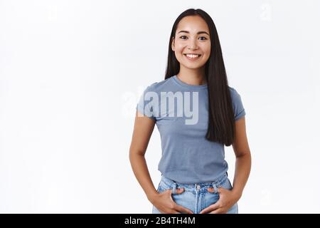 Fröhliche, unbeschwerte junge asiatische Freundin mit lond dunkler Frisur, hält die Hände in den Jeans-Taschen und lächelt froh, sieht kamerabegeistert aus Stockfoto