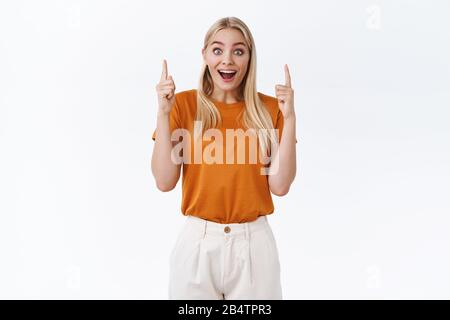 Begeisterte, gut aussehende blonde Frau in orangefarbenem T-Shirt, Tätowierungen an den Armen, lächelnd gewundert und amüsiert, sehen überrascht aus, zeigen die Finger staunen Stockfoto