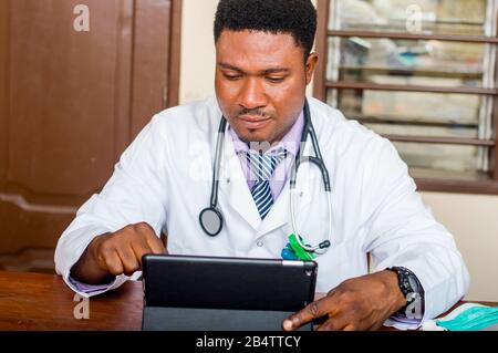 Junger männlicher Arzt sitzt am Tisch und arbeitet an seiner Tablette in seinem Büro im Krankenhaus. Stockfoto