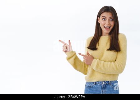 Großartige Neuigkeiten, die Sie sich ansehen können. Aufgeregt glücklich und überrascht schöne junge Frau im gelben Pullover Aufmerksamkeit auf Banner, Zeigefinger nach links und Stockfoto