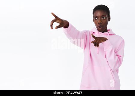 Faszinierte und neugierige junge afroamerikanische stylische Frau in rosafarbenem Hoodie, die seitlich zeigt und sieht etwas Interessantes auf der Distanz Stockfoto