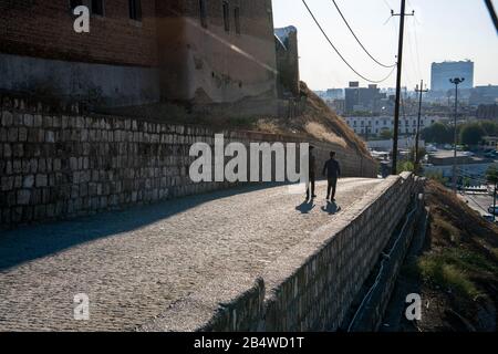 Irak, Iraq Kurdistan, Arbil, Erbil. Zwei Männer gehen auf einer Straße spazieren. Auf der linken Seite befindet sich die Zitadelle Qalat. Im Hintergrund ein Blick auf Erbil bei Sonnenuntergang. Stockfoto