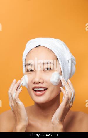 Lachende Mädchen Anwendung Feuchtigkeitscreme auf ihrem Gesicht. Foto der jungen Mädchen mit makelloser Haut auf orangem Hintergrund. Hautpflege und Schönheit Konzept Stockfoto