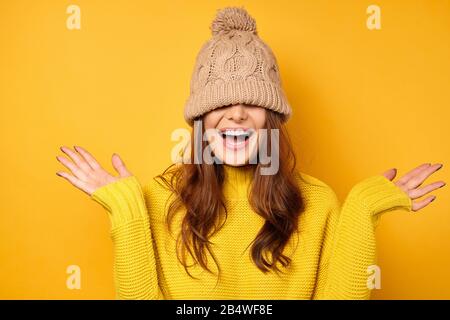 Ein Mädchen in einem gelben Pullover steht auf einem gelben Hintergrund mit einer Kappe, die über das Gesicht gezogen wird, und lacht und verteilt die Arme nach außen zu den Seiten Stockfoto