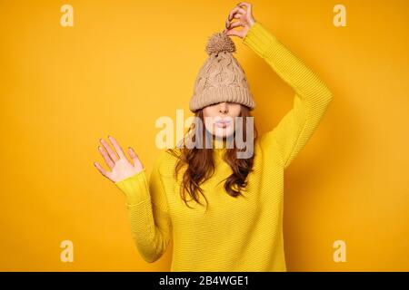 Eine Brunette in einem gelben Pullover steht auf gelbem Grund mit einer über ihr Gesicht gezogenen Kappe und zieht sich für einen Ponpon nach oben Stockfoto