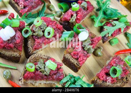 Herzhaft heftiger deutscher Imbiss: Isolierte Holzplatte mit geröstetem tataren-knoblauchbrot, Frühlingszwiebeln, Kapern und Rucola Salatblättern Stockfoto