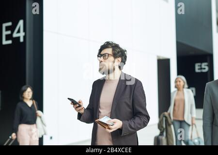 Horizontales mittleres Porträt des nachdenklichen jungen Mannes, der in der Flughafenhalle steht und Smartphone, Bordkarte und Geldbörse mit Blick auf die Informationsanzeige hält Stockfoto