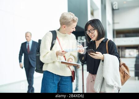 Zwei glückliche junge Frauen, die mit ihrem Gepäck am Flughafen stehen, und Dokumente, die auf ihren Flug warten, horizontaler Schuss Stockfoto