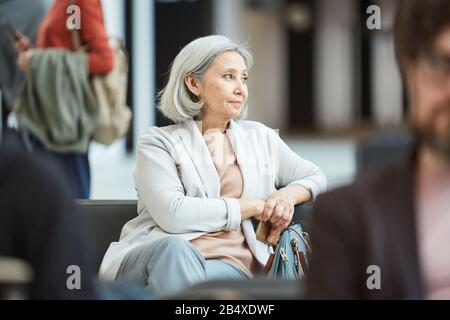 Horizontales Porträt reifer Frau mit grauem Haar entspannt in der Abflughalle sitzend, wegblickend an etwas denken, Kopieraum Stockfoto