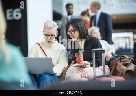 Zwei junge weibliche Freunde sitzen zusammen in der Abflughalle und beobachten etwas Lustiges auf dem Laptop, horizontales Porträt Stockfoto