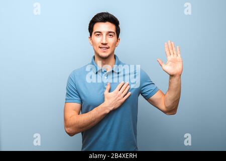 Ansehnlicher junger Mann mit neutralem Lächeln in blauem Poloshirt mit Hand auf der Brust, der Eid vor blauem Hintergrund abgibt. Studio Shoot Stockfoto