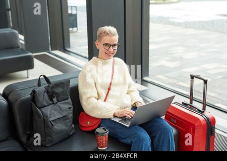 Horizontaler Hochwinkelschuss von stilvoller blonder Frau, die Brille in der Abflughalle trägt, mit ihrem Laptop mit Blick auf die Kamera Stockfoto