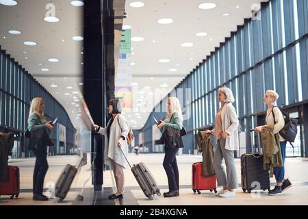 Horizontaler Seitenansicht von vier Frauen, die in der Warteschlange mit ihrem Gepäck am Check-in-Schalter des Flughafens stehen, Kopierraum Stockfoto