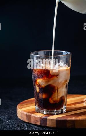 Milch, Die in Eiskaffee in Hochgläserem Glas auf dunklem Hintergrund Gegossen wird. Konzept Erfrischendes Sommergetränk. Stockfoto