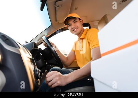Glücklicher Liefermann in gelber Poloshirt-Uniform mit Paketkarton im Auto. Lächelnder Mann der Kurierzustellung, der im Auto sitzt und den Wahnvorstellung überprüft Stockfoto