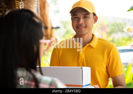 Glücklicher Liefermann in gelber Poloshirtuniform mit Paketpostbox in den Händen vor den Haustüren des Kunden. Lächelnde Kurierzustellung mA Stockfoto