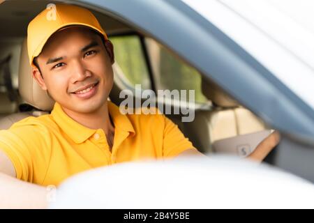Glücklicher Liefermann in gelber Poloshirt-Uniform mit Paketkarton im Auto. Lächelnder Mann der Kurierzustellung, der im Auto sitzt und den Wahnvorstellung überprüft Stockfoto